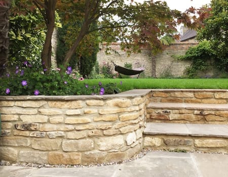 A hammock sits on the raised lawn under an acer tree and surrounded by bright purple Geranium Rozanne's